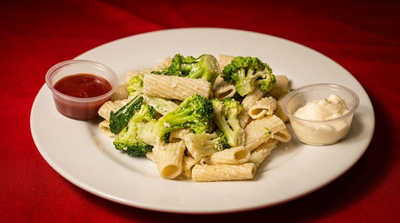 Rigatoni pasta with broccoli and creamy sauce, served with ketchup and mayonnaise on a white plate.