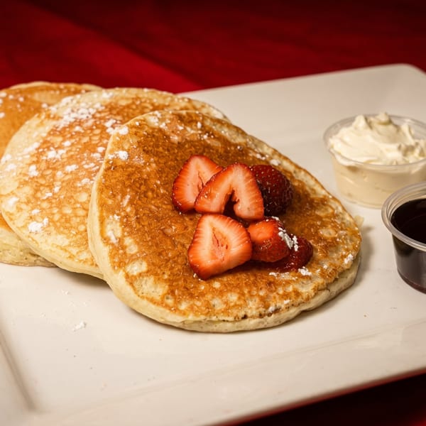 Fluffy pancakes topped with strawberries and powdered sugar, served with syrup and whipped cream.